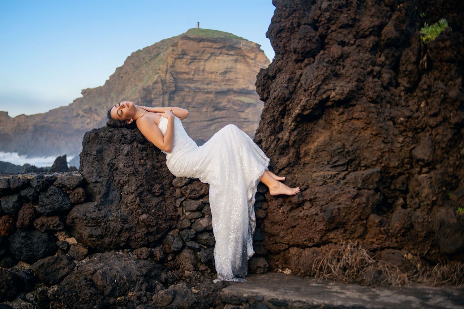 A figure in a flowing white dress poses on volcanic rocks with ocean cliffs in the background.