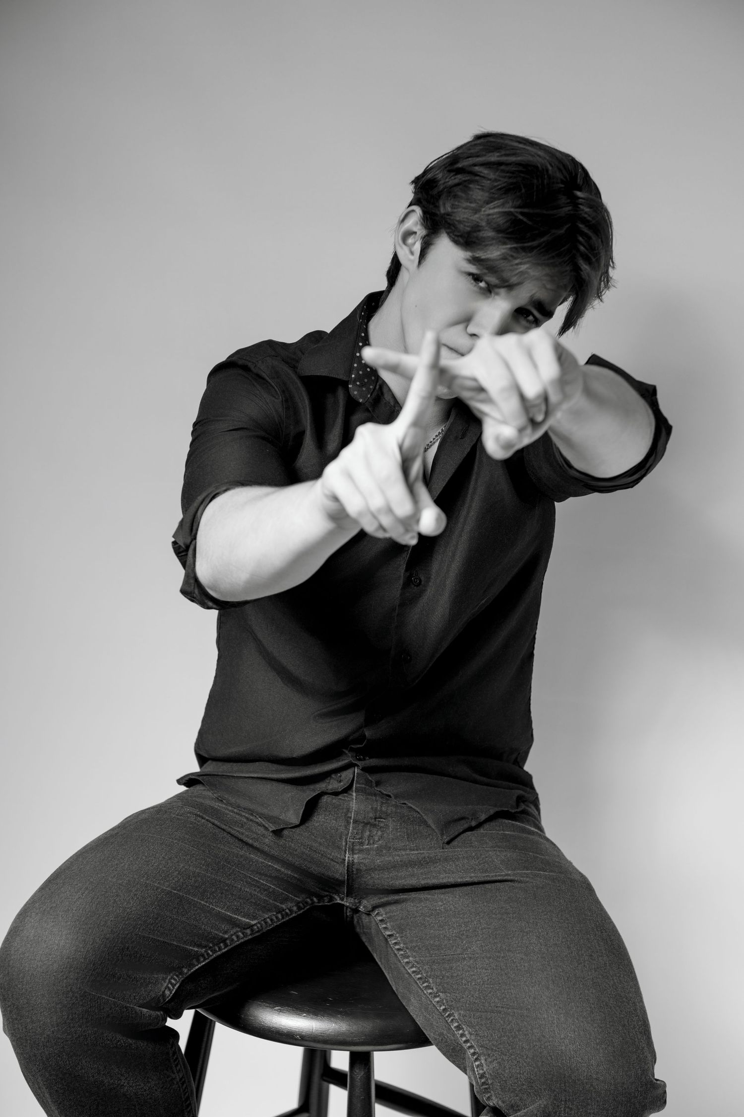 Black and white portrait of person making hand gestures while seated on a stool.