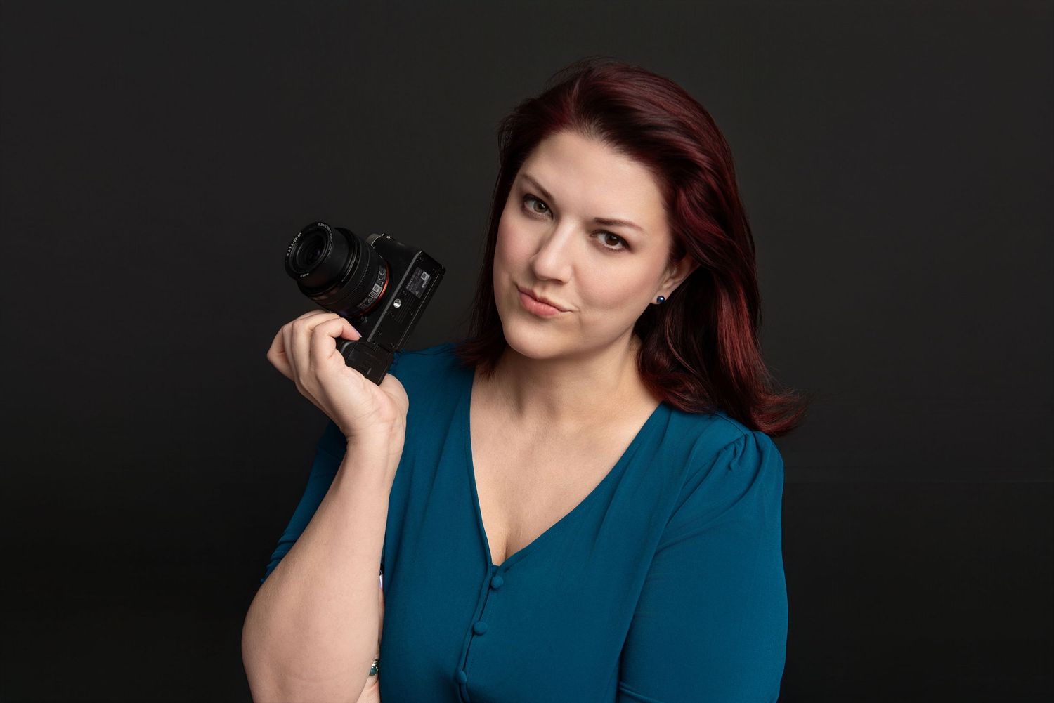 A woman with red hair in a teal dress holds a camera, posing confidently against a dark background.