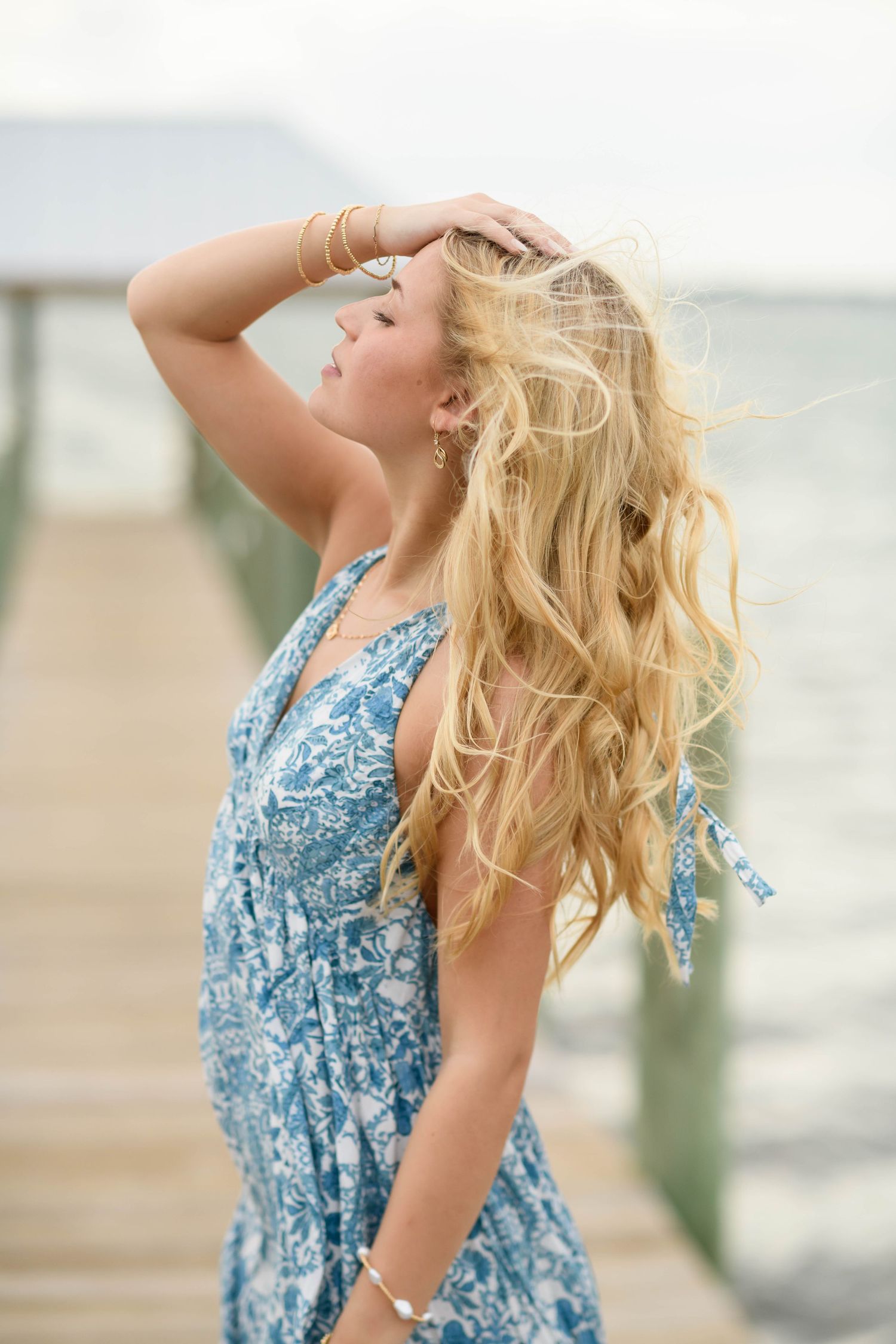 Woman in blue floral dress with flowing hair poses on wooden pier by ocean.