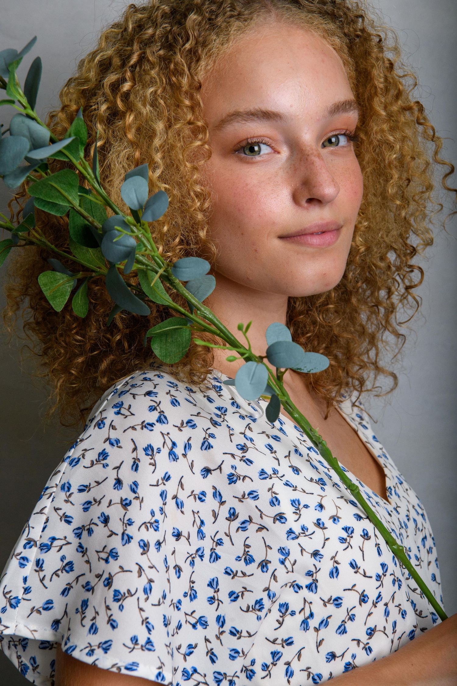 Close-up portrait wearing blue floral print dress with eucalyptus branch against grey background.