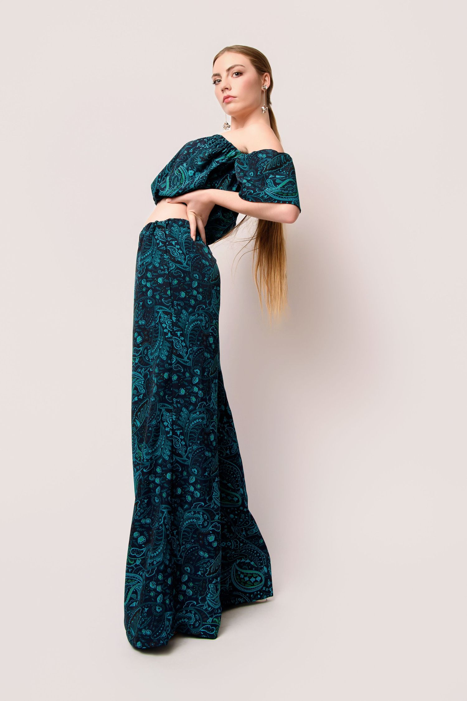 Model wearing teal floral maxi dress poses elegantly against light backdrop.