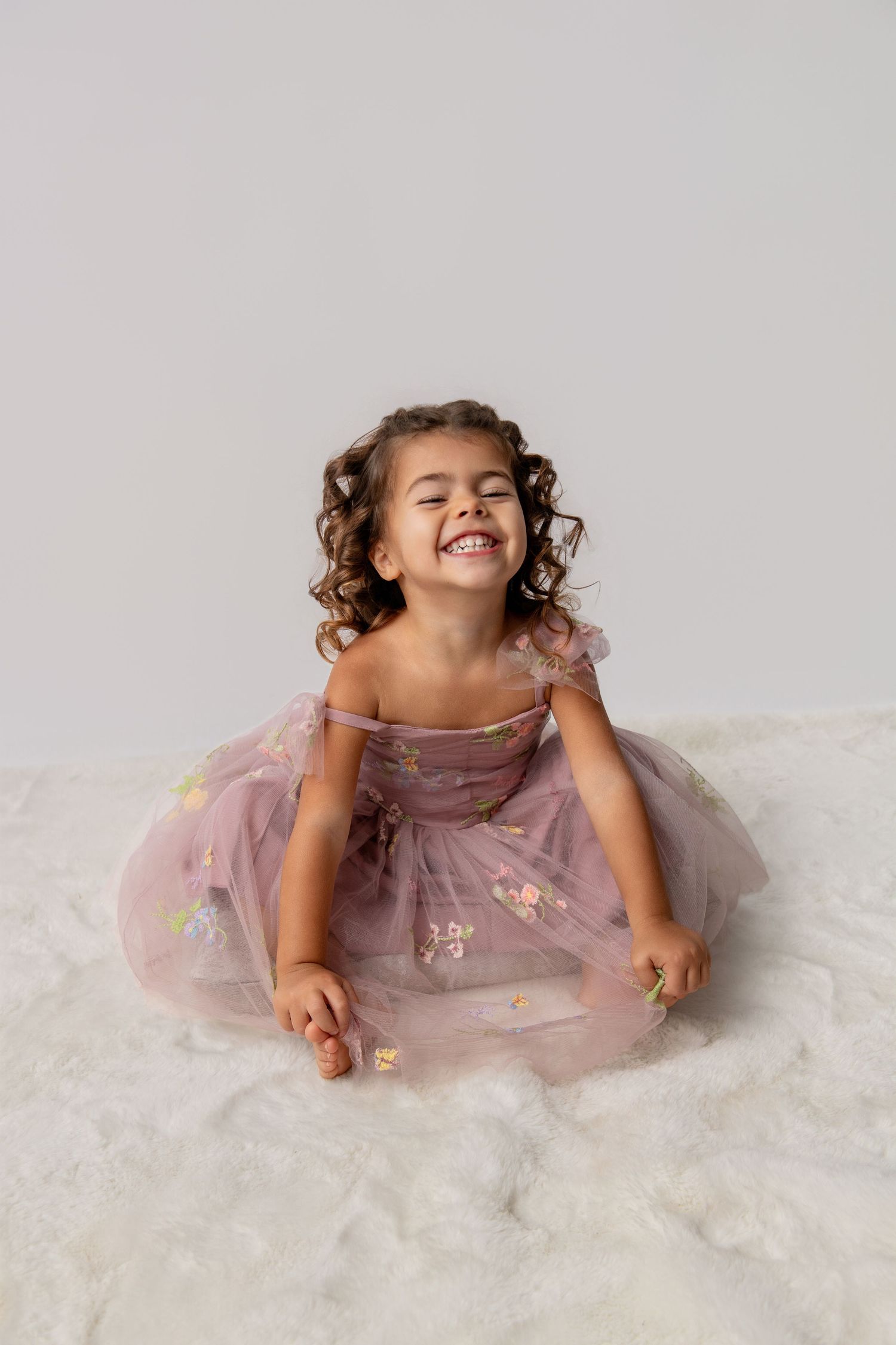 Young child in purple tulle dress laughing and playing on white backdrop.