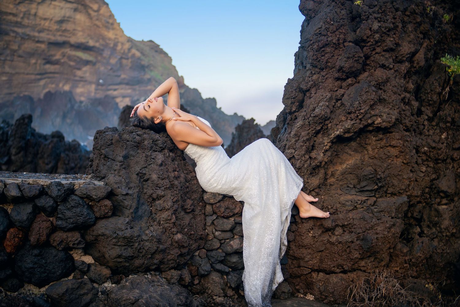 A figure in a flowing white dress reclines dramatically on dark volcanic rocks against a misty coastal backdrop.