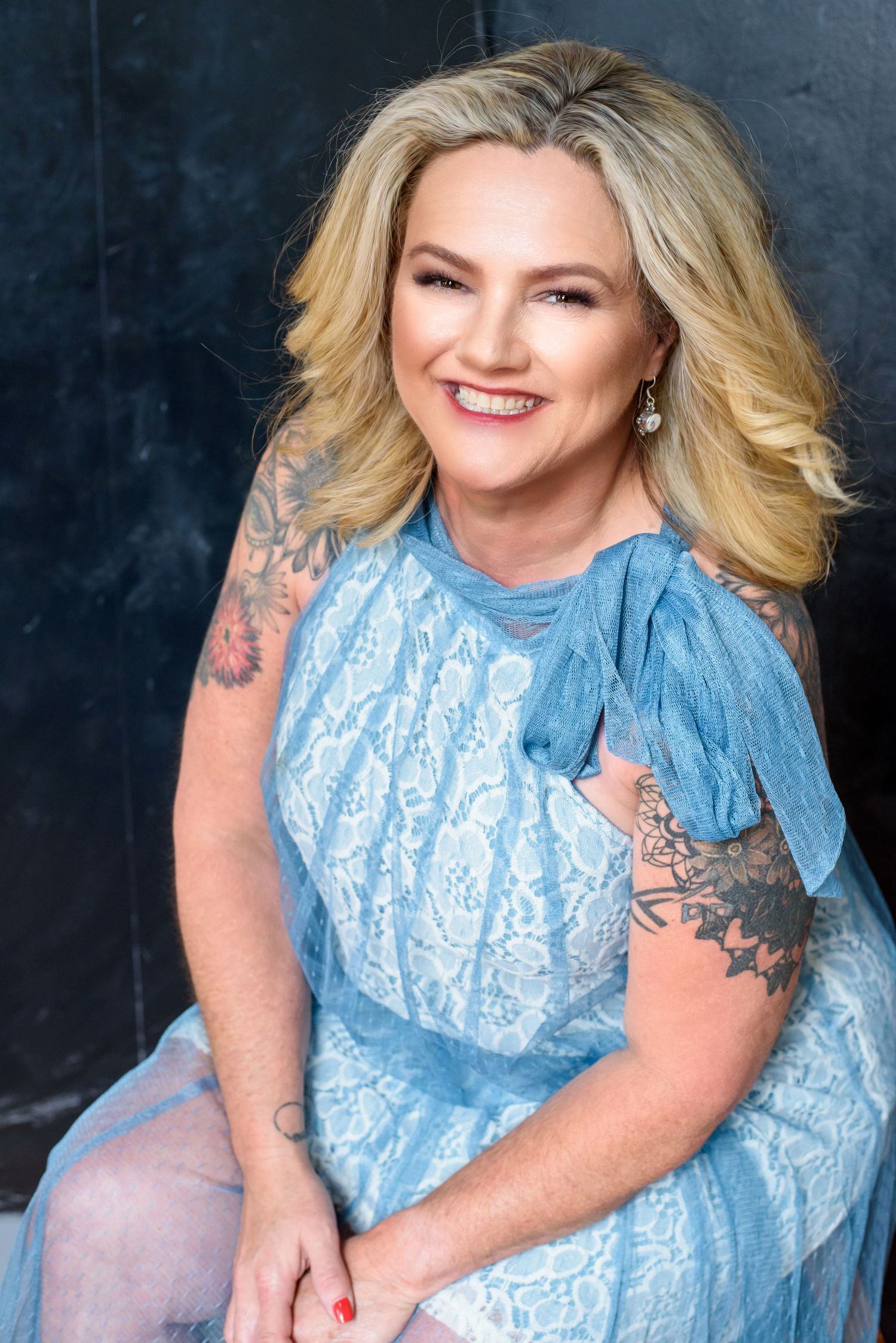 Joyful portrait in a light blue lace dress with visible arm tattoos and blonde hair.