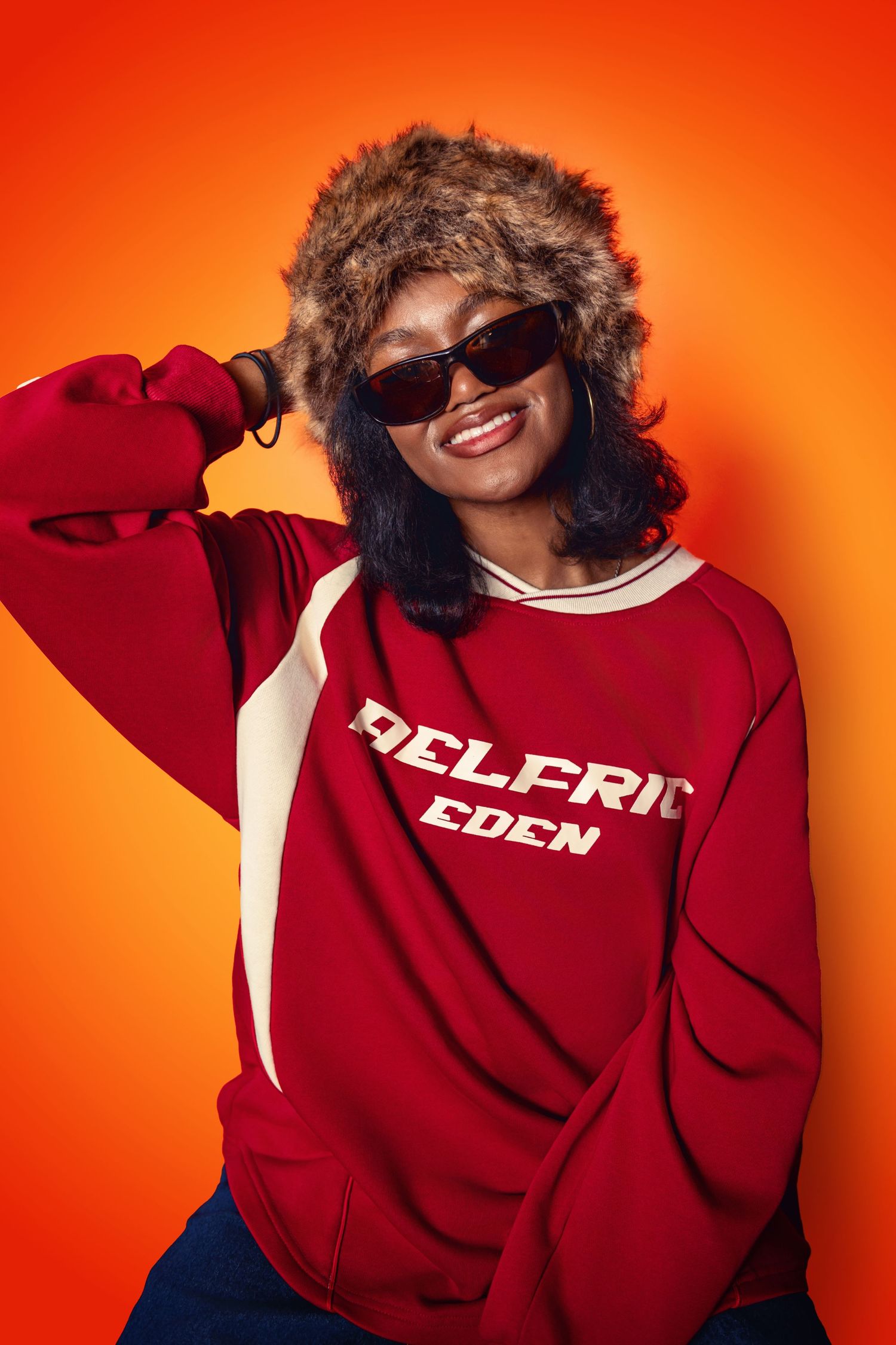 Person in red sweatshirt and fur-trimmed hood poses playfully against orange background.