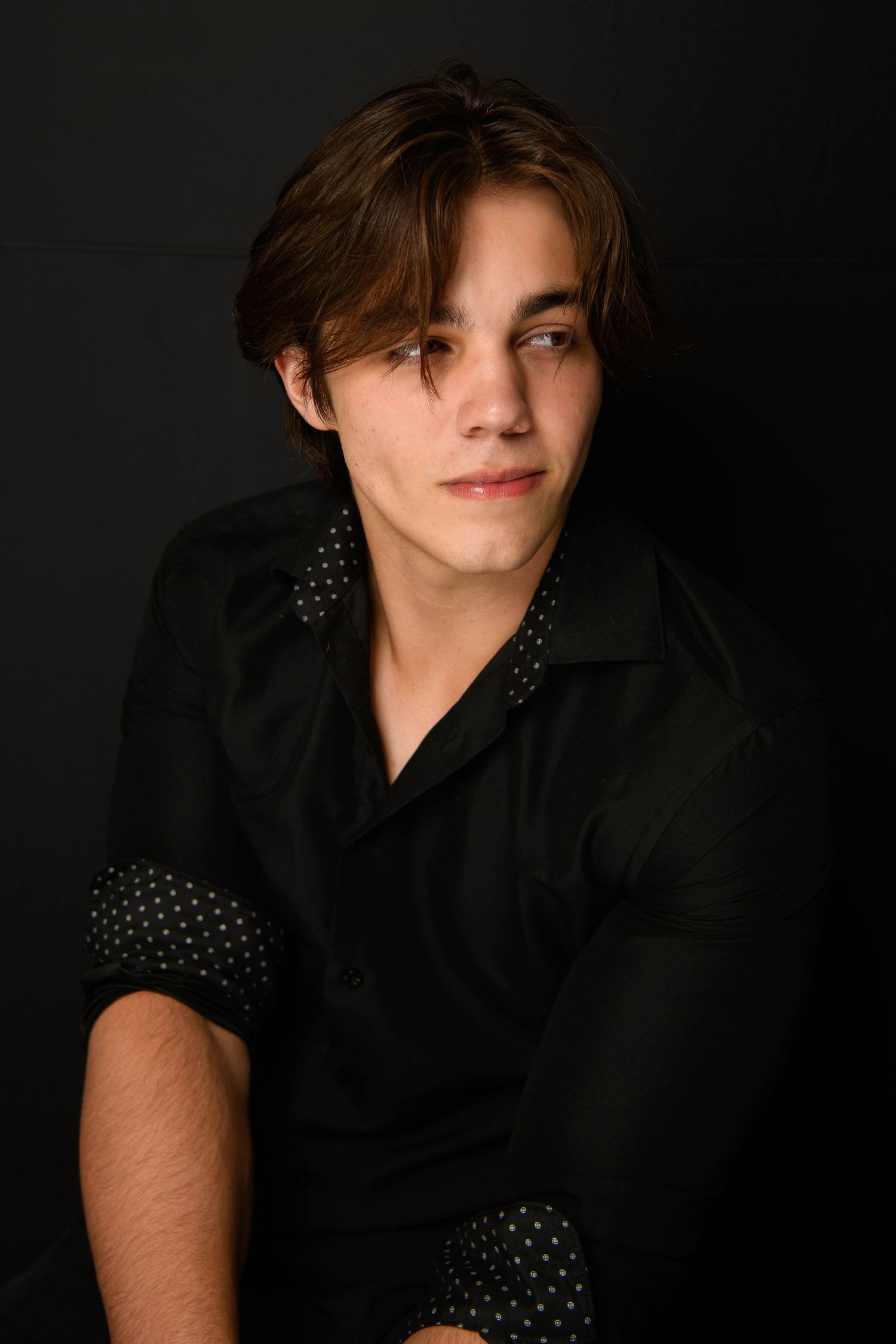 Young man with brown hair wearing a black polka dot shirt, posed against a dark background in a moody portrait.