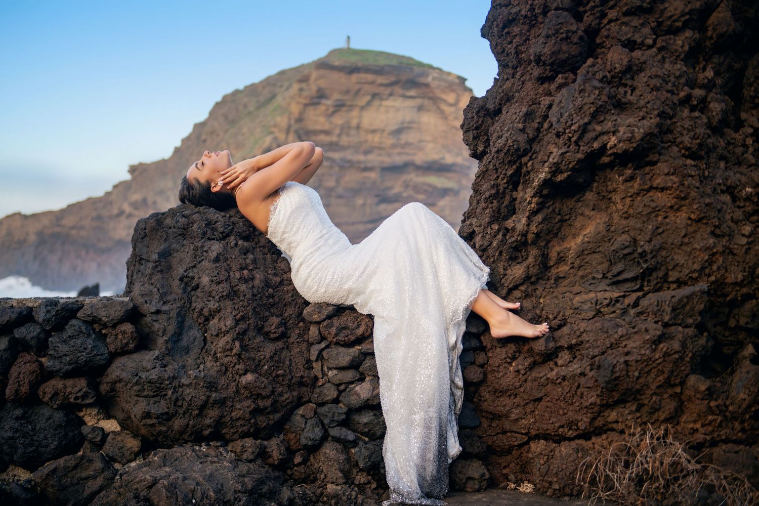 A person in white fabric reclines on dark coastal rocks with mountains and ocean in the distance.