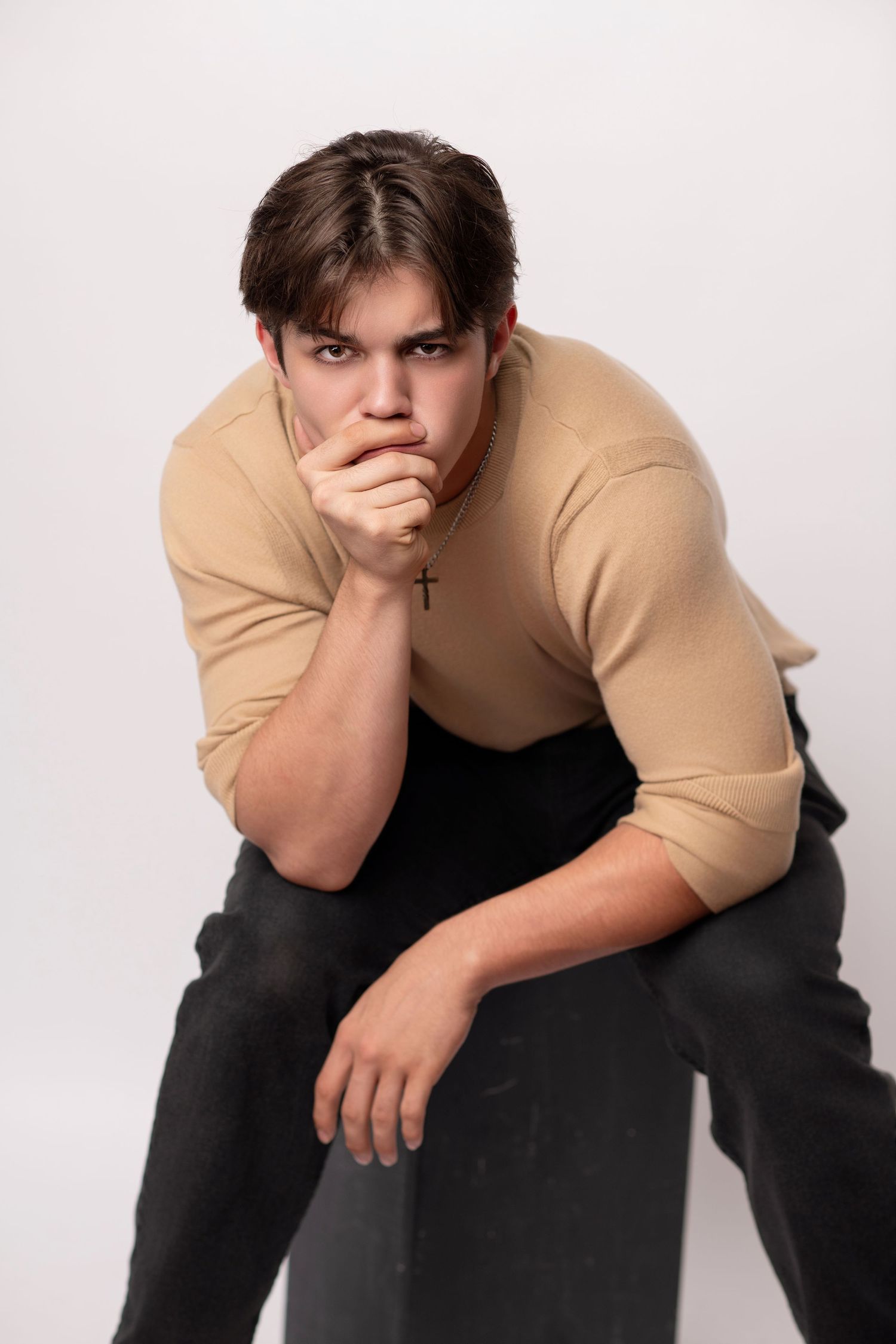 Studio portrait of person in beige sweater and dark pants in contemplative pose.