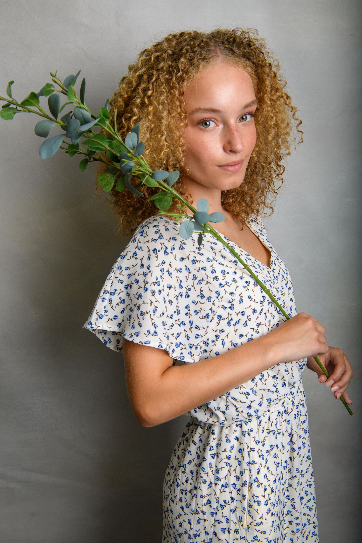 Portrait with curly hair wearing white and blue floral dress against grey background with greenery.