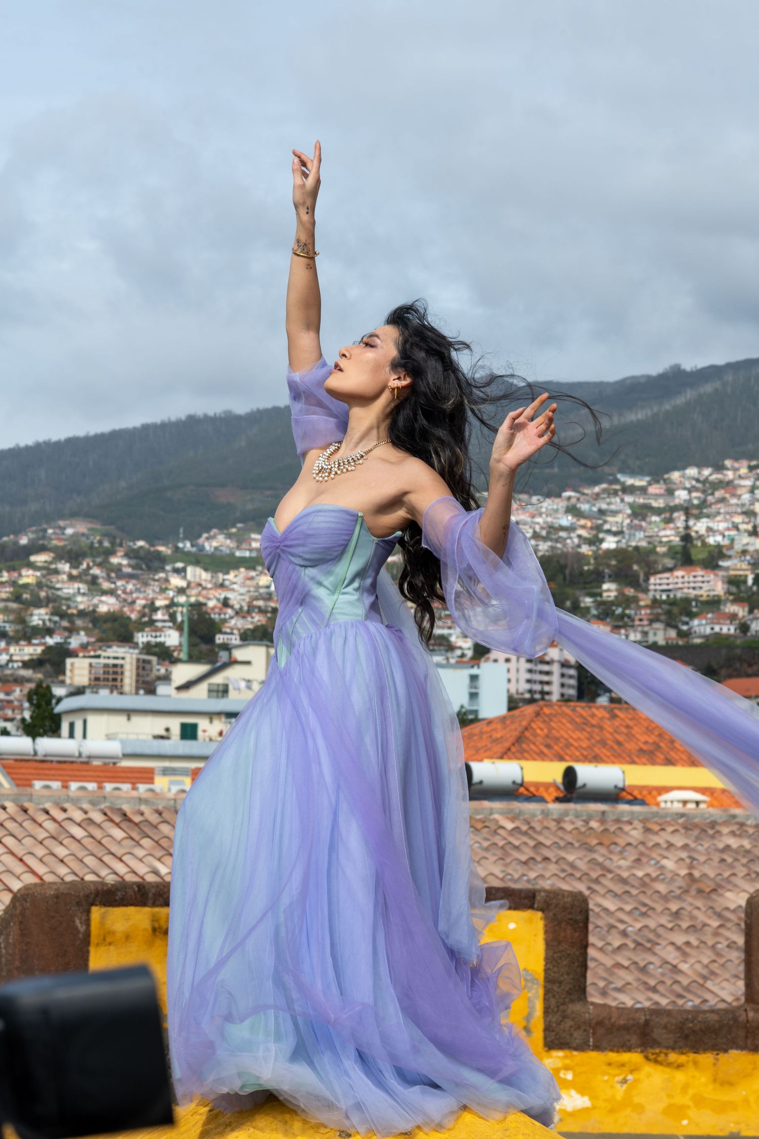 Dancer in lavender gown performs expressive movements on rooftop with city backdrop.
