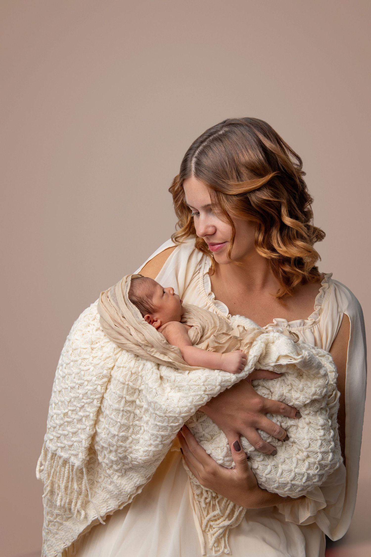 Mother in white dress holding newborn baby in cream blanket against neutral background.