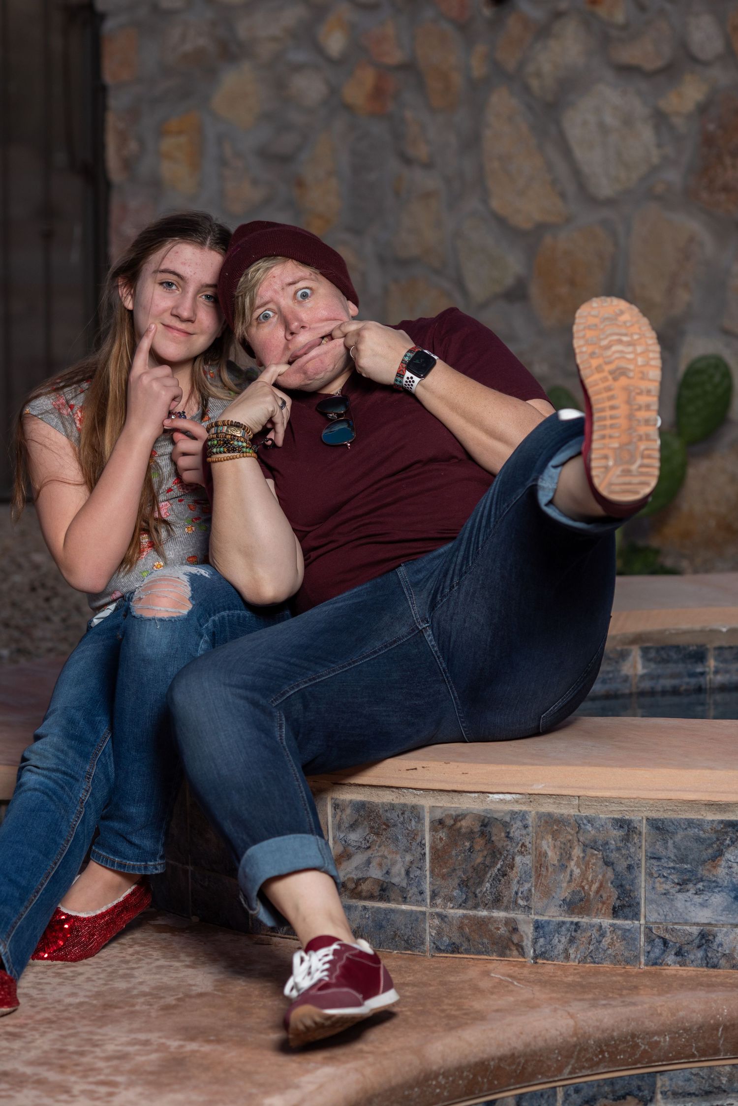 A playful couple lounges on a stone ledge in casual clothing against a rustic stone wall background.