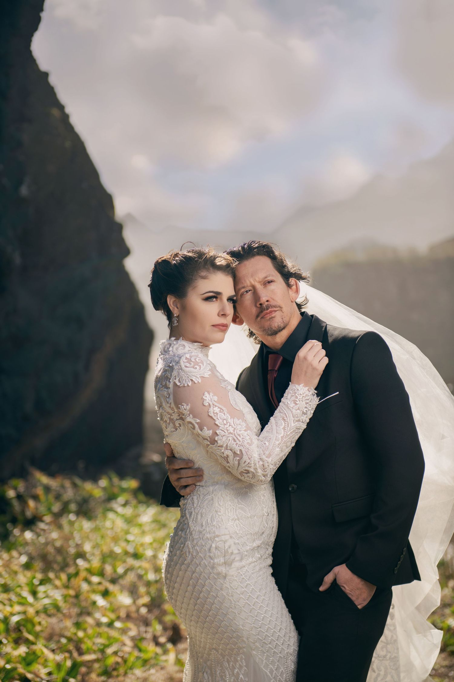 Bride in lace gown and groom share tender moment against scenic backdrop.