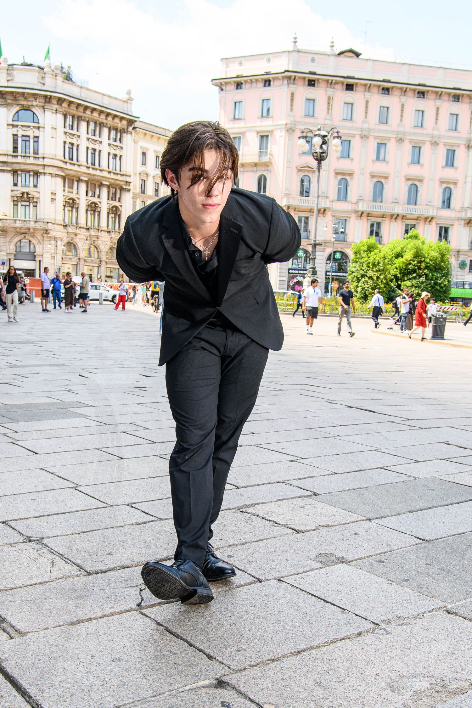 Stylish figure in all black suit striking dynamic pose in Milan city square with historic architecture.