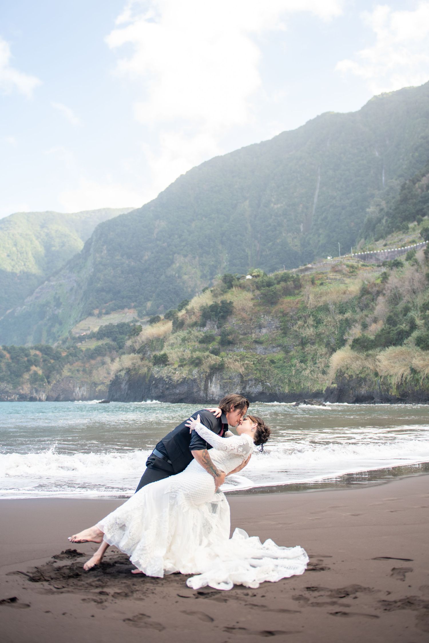 A couple poses dramatically on a black sand beach with mountains and ocean waves in the background.