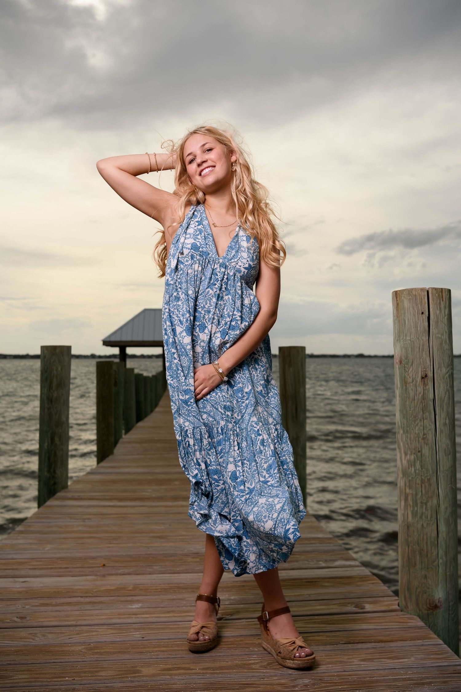 Woman in flowing blue patterned dress poses on wooden dock at sunset.