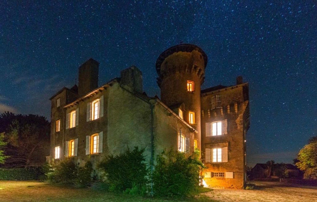 Historic stone castle illuminated at night under a stunning starry sky, with glowing windows and a round tower.