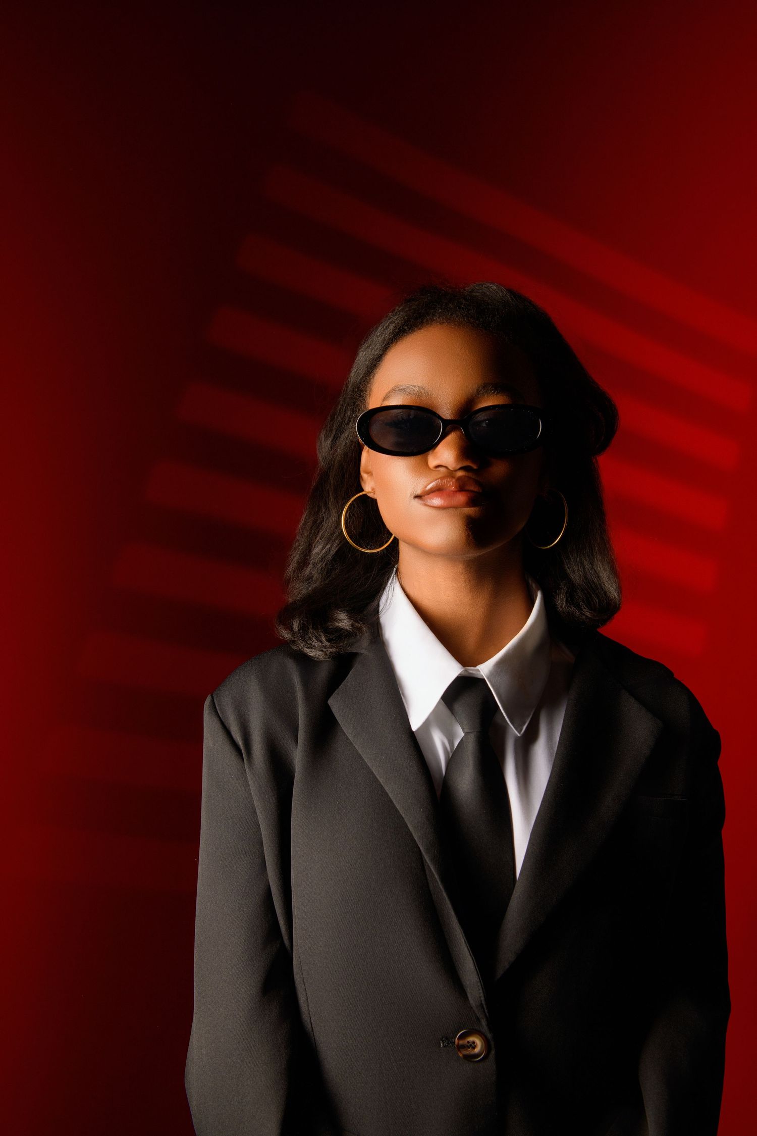Fashion model in black suit and sunglasses poses mysteriously against red backdrop.