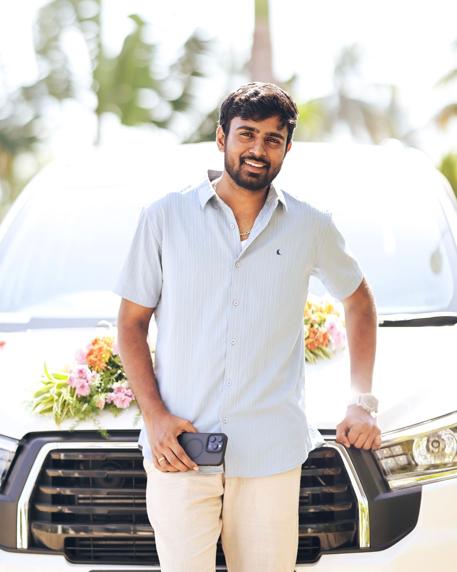 Man in casual white outfit standing near a luxury car on a bright sunny day.