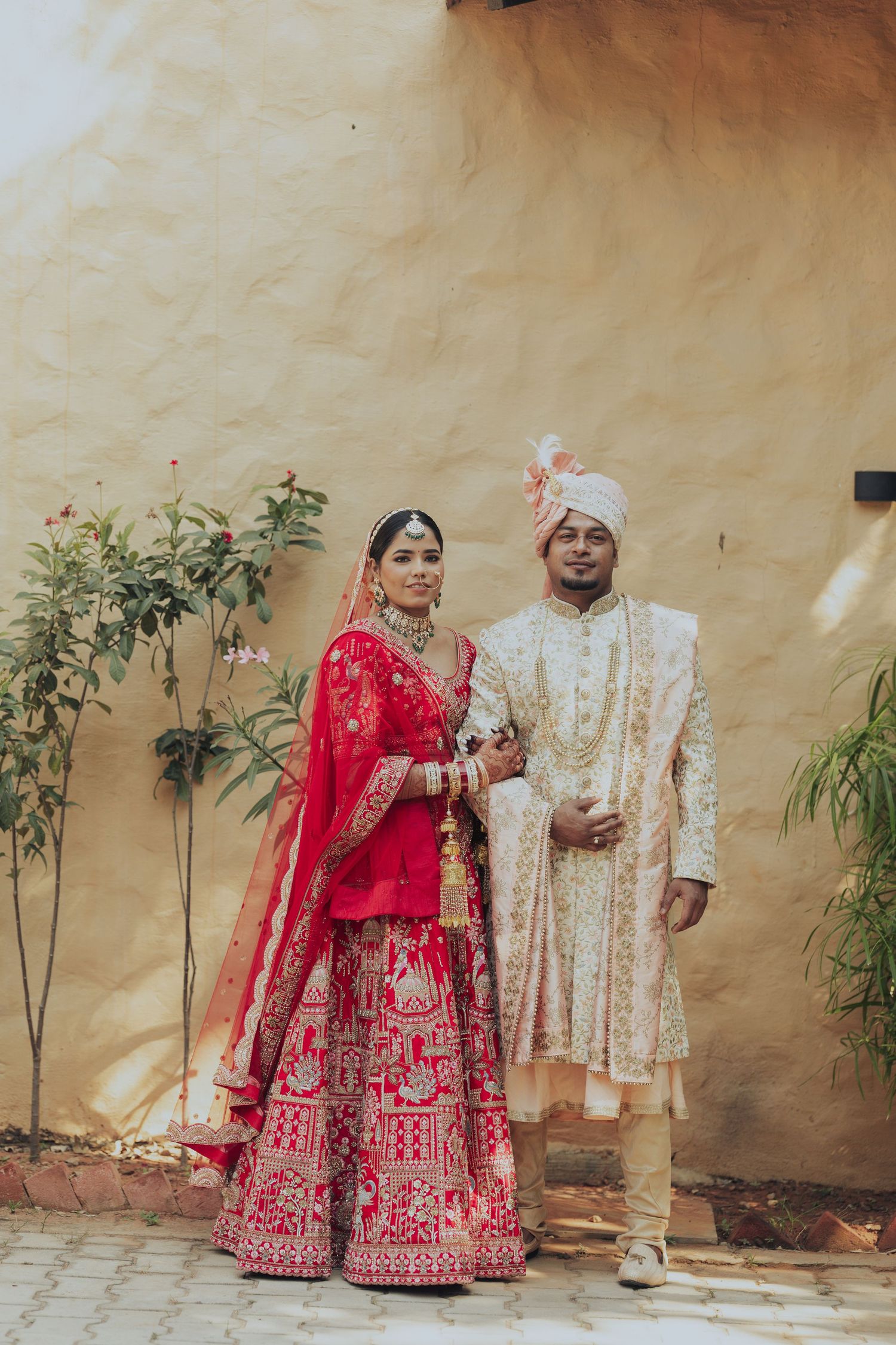 Traditional Indian wedding couple in red and cream outfits against beige wall with plants.