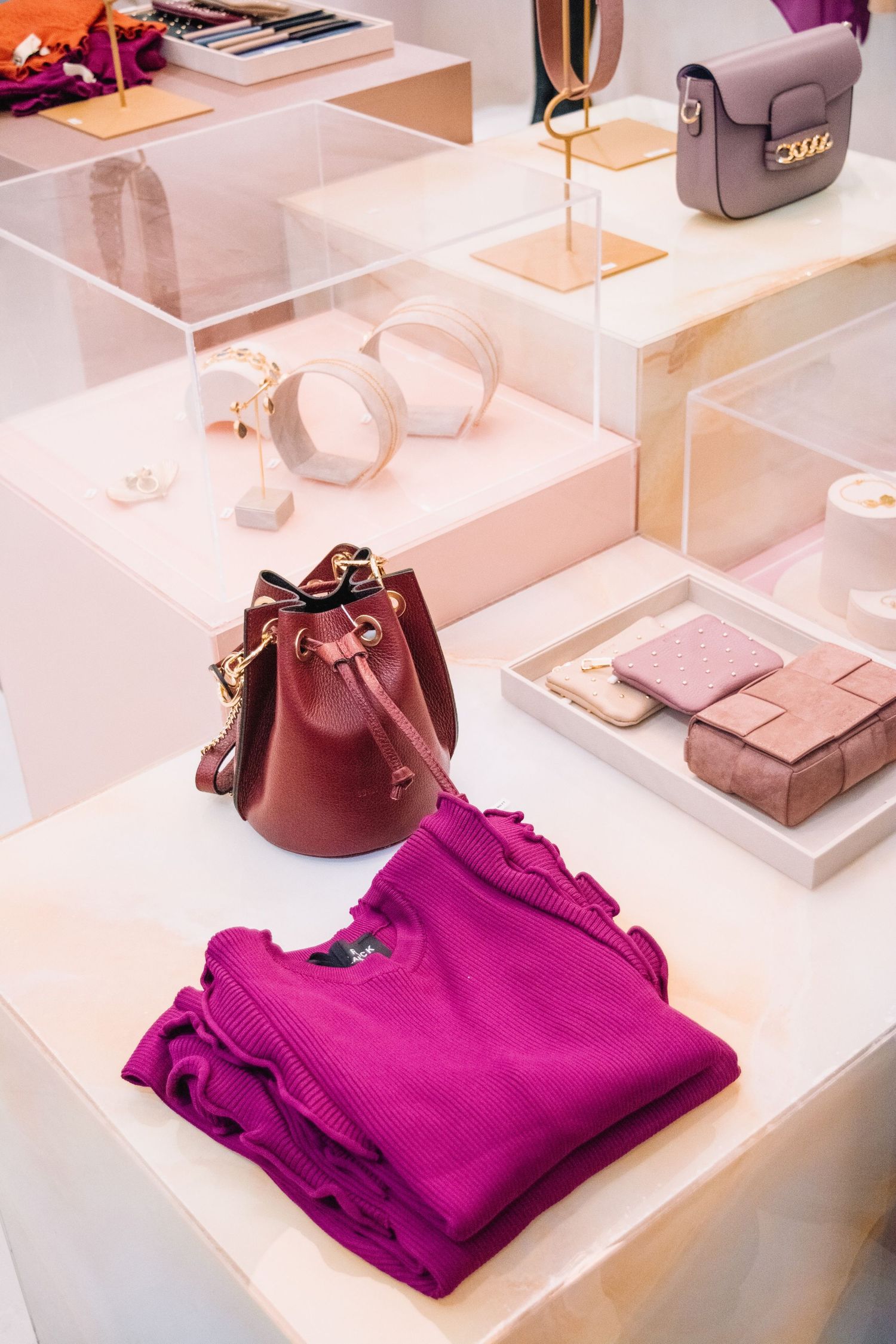 Luxury boutique display with magenta knit sweater, burgundy bucket bag, and accessories on pink shelving.