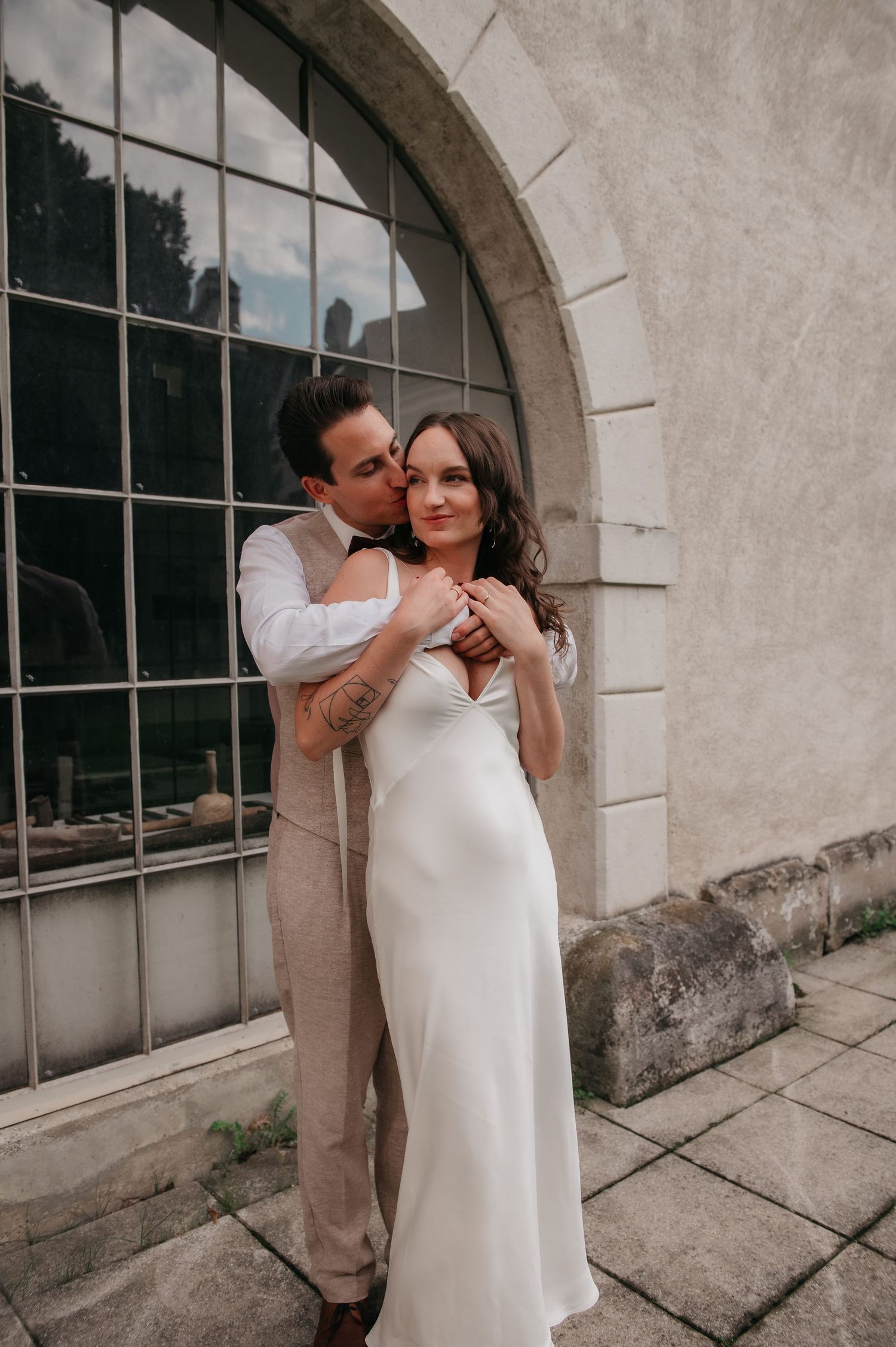 Groom kissing bride on cheek while embracing her in front of arched window in elegant wedding attire.