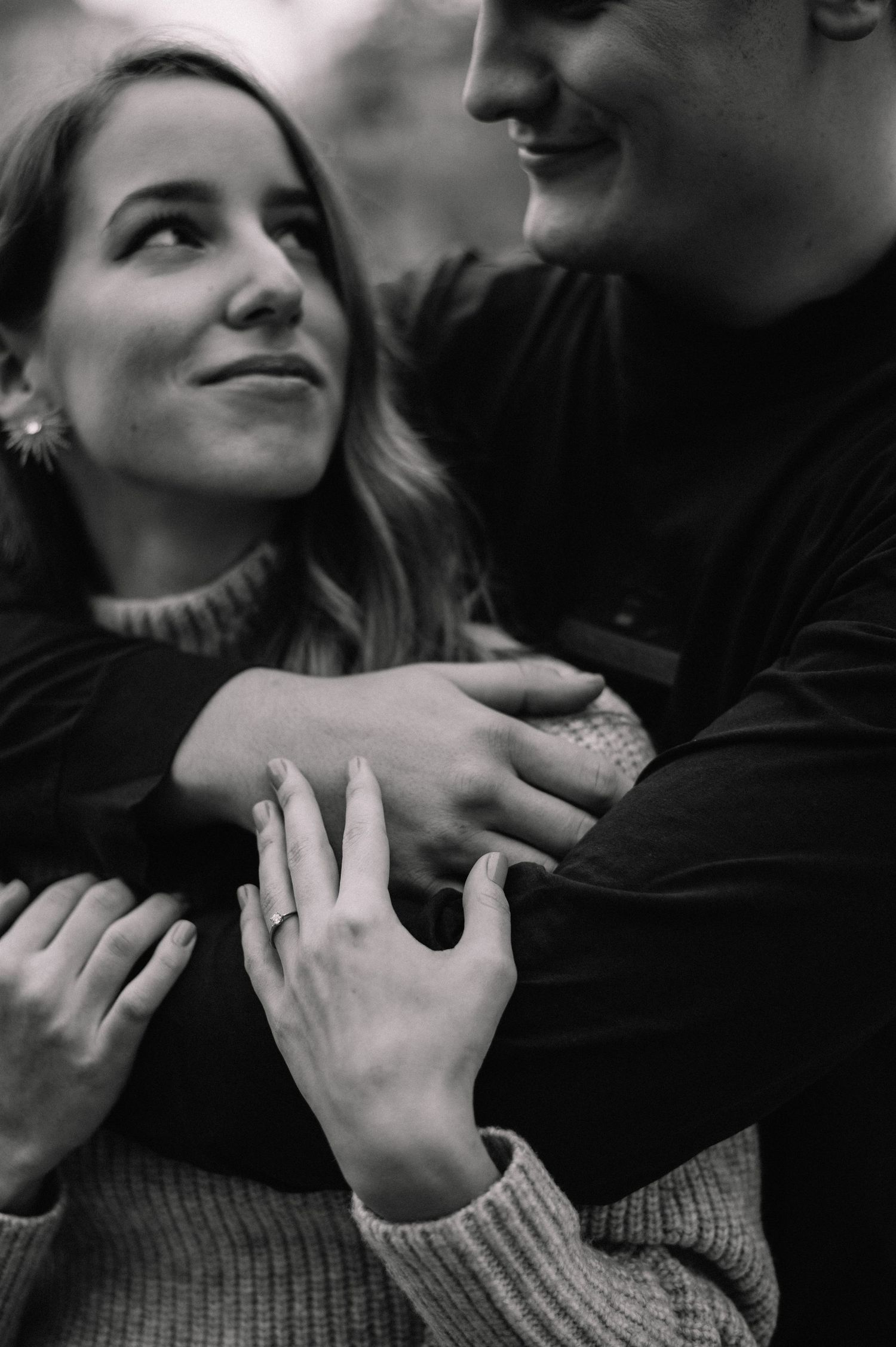 Black and white photo of a couple embracing, woman gazing up lovingly, engagement ring visible on her hand.
