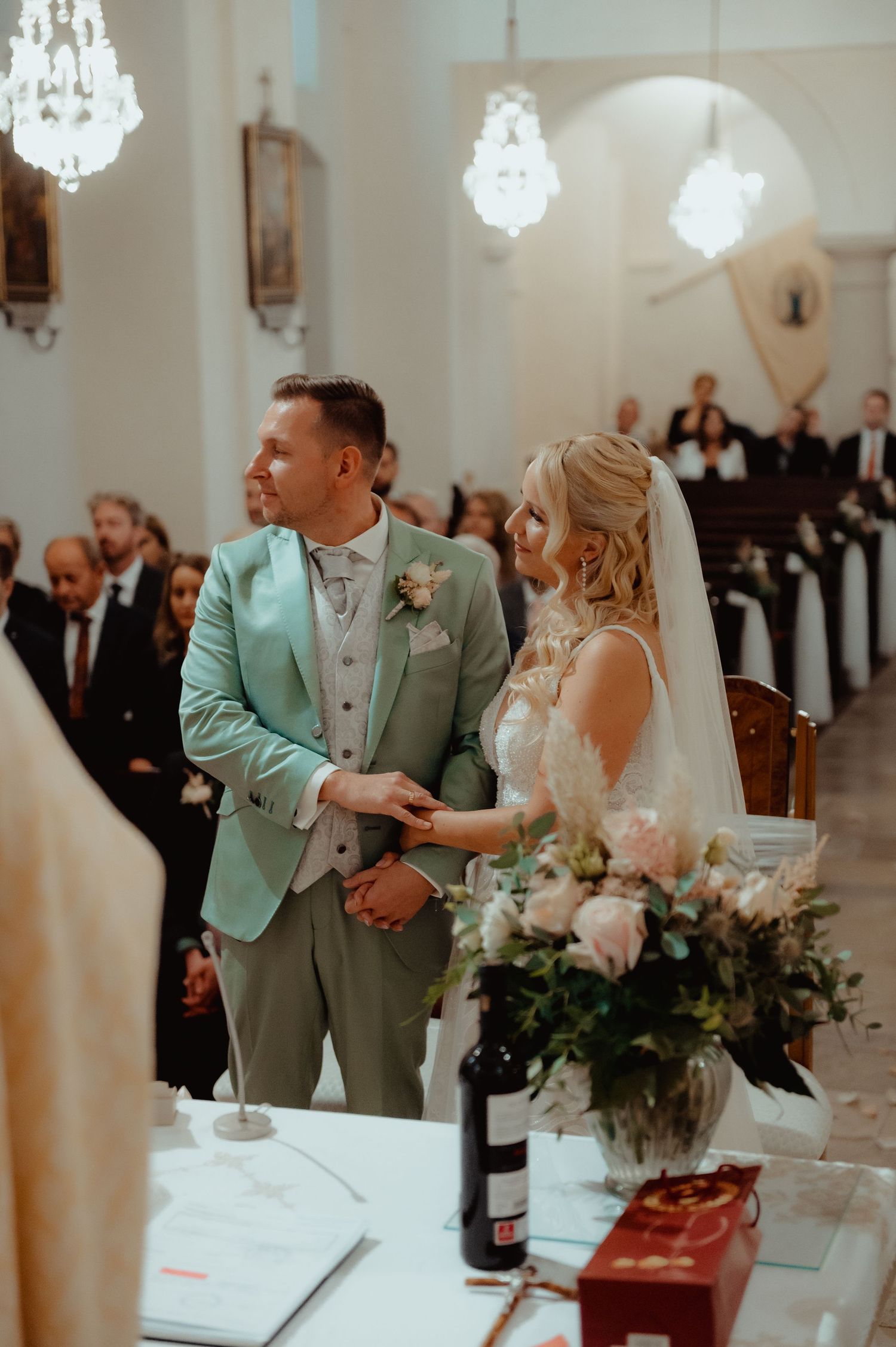 Bride and groom holding hands at church altar during wedding ceremony, groom in mint suit, bride in lace gown.