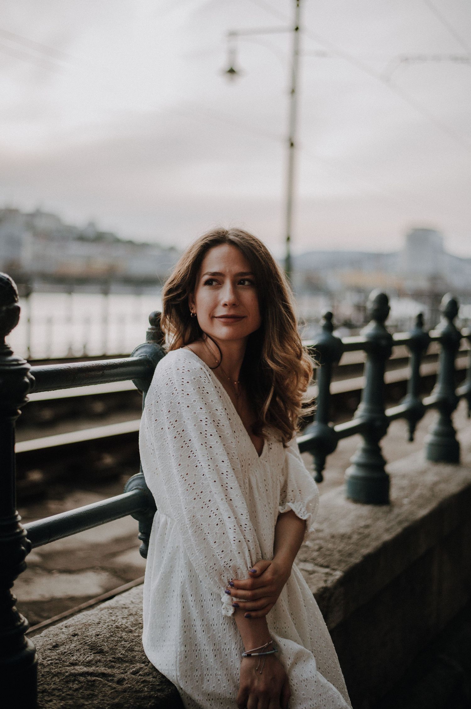 Woman in white eyelet dress sitting by ornate iron railing near waterfront at dusk.
