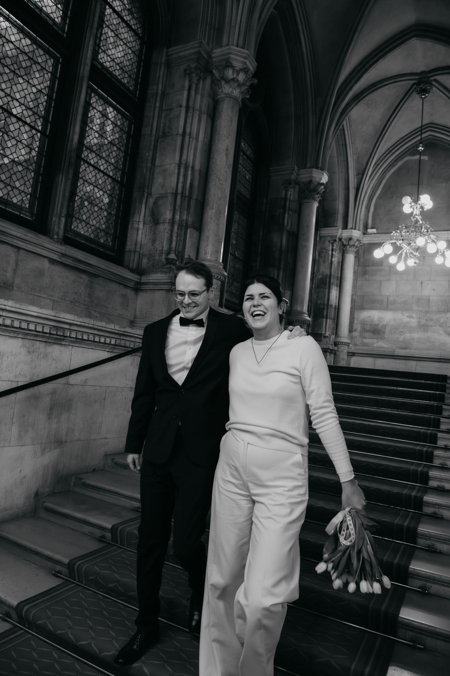 Black and white photo of a joyful couple in wedding attire descending grand stone stairs in an elegant historic building.