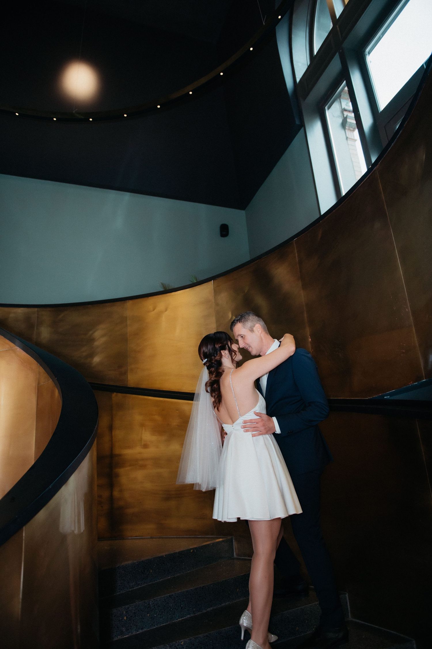 Bride in short white dress and groom embrace on a curved golden staircase in a modern venue.