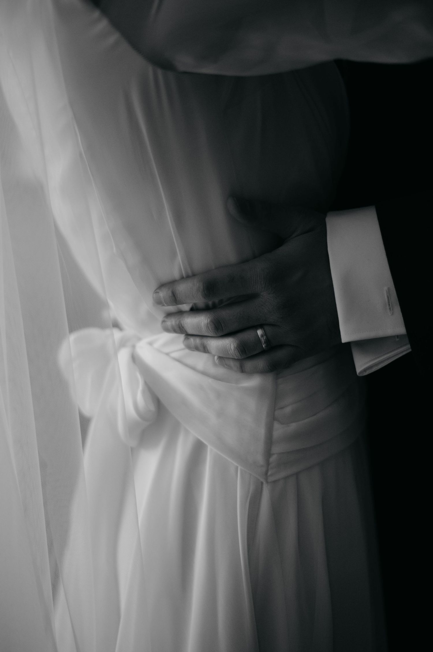 Black and white close-up of grooms hand with ring holding brides waist in white gown.