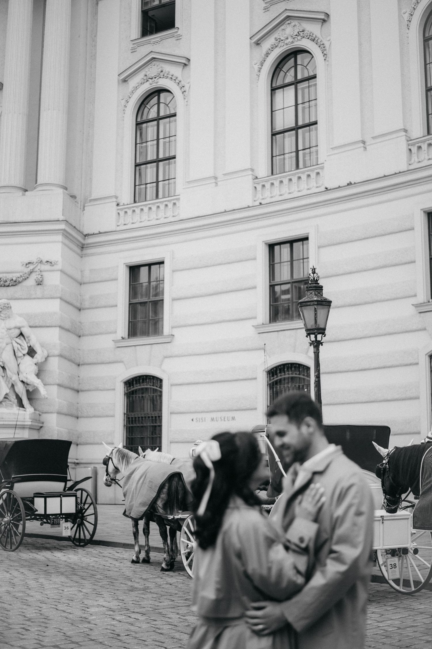 Black and white photo of a couple embracing in front of a grand European building with horse-drawn carriages in Vienna.