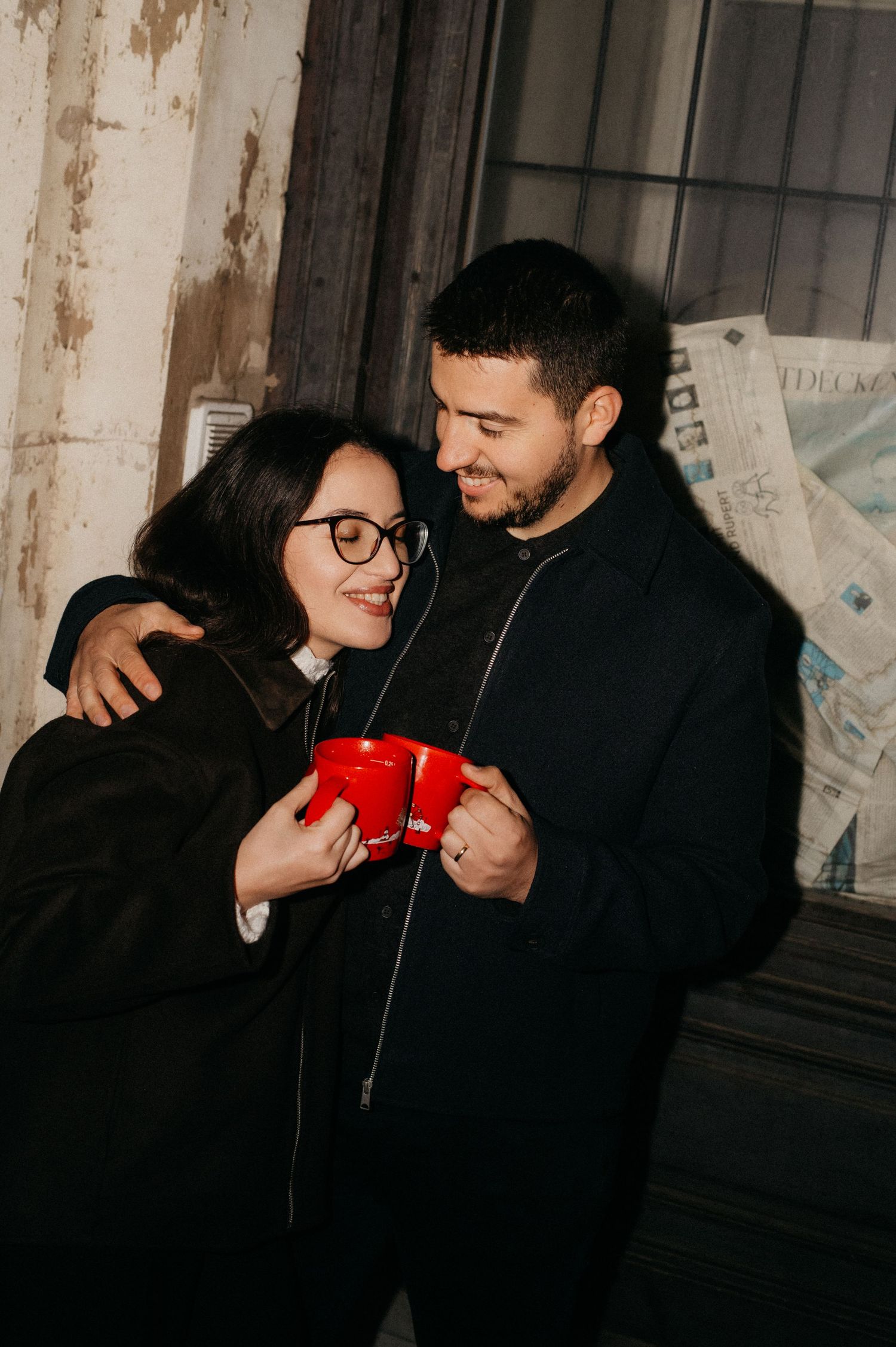 Couple in black coats sharing red mugs, smiling and embracing near a rustic wall with newspapers.