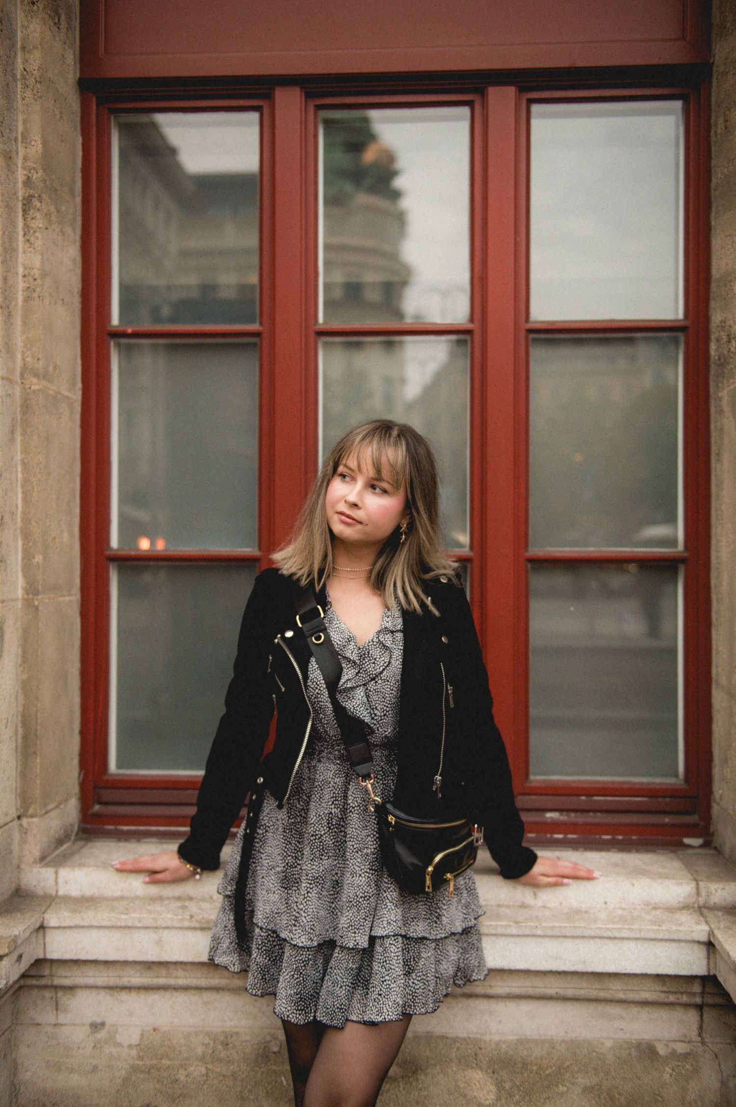 Young woman in black moto jacket and gray ruffle dress with belt bag poses before red-framed windows.
