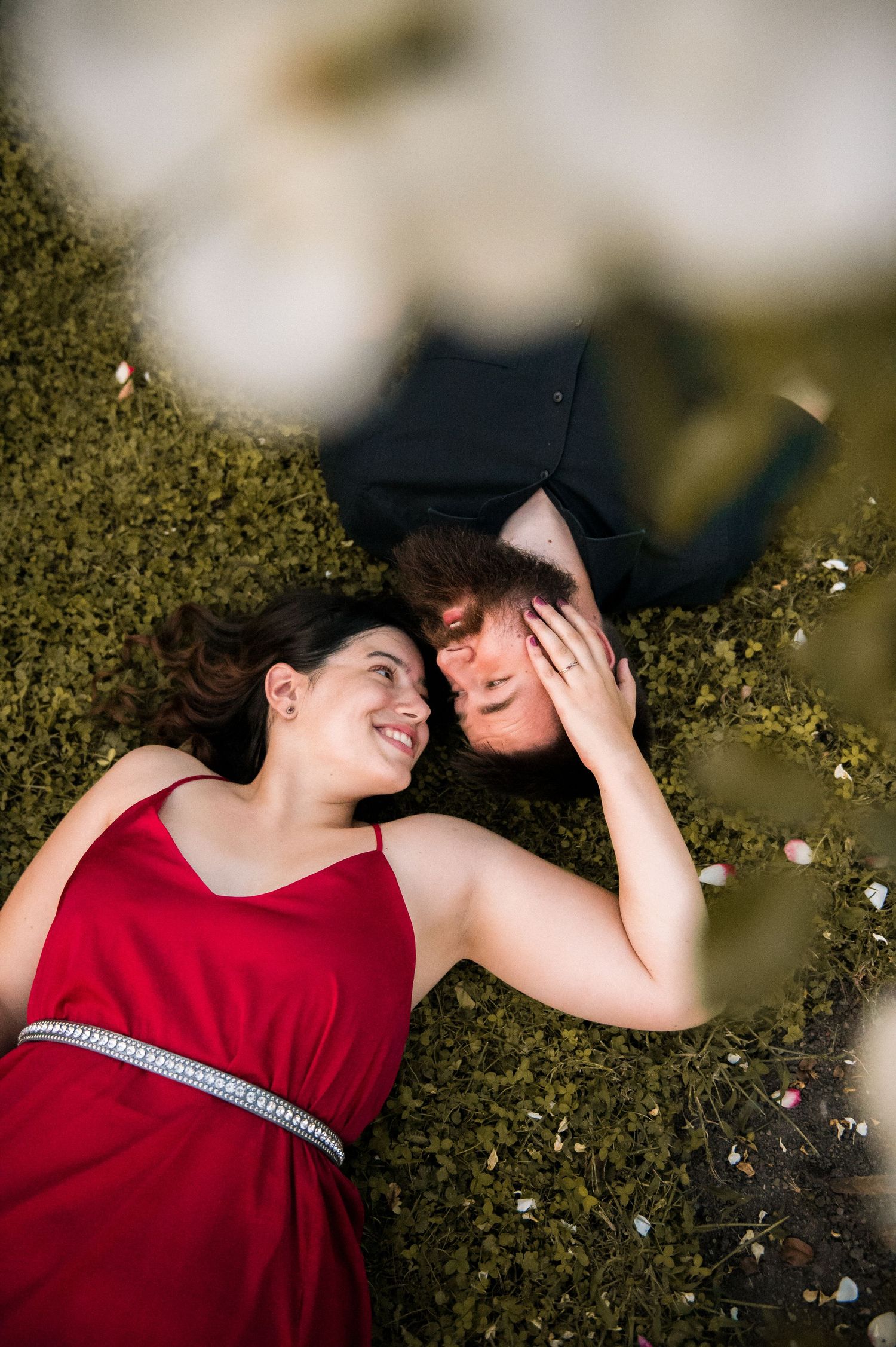 Couple in red dress and graduation cap lying on grass, smiling at each other in a romantic outdoor setting.