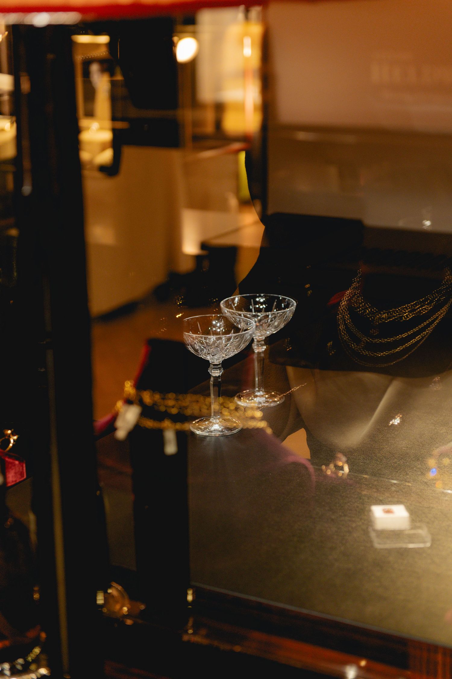 Two elegant crystal coupe glasses displayed on a dark surface in a warmly lit jewelry store showcase.
