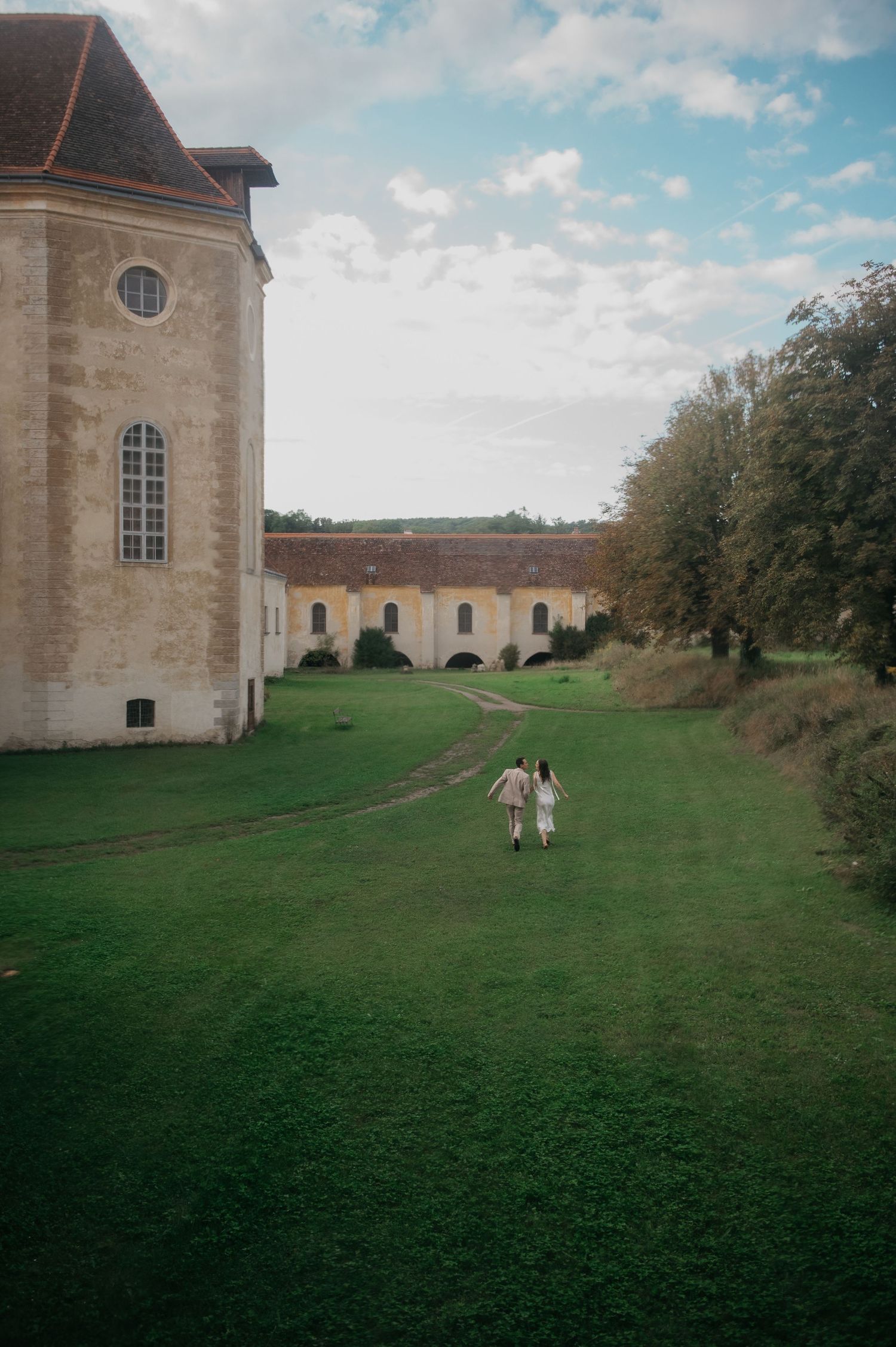 Two people walk hand-in-hand across lush green grounds beside a historic European estate under a partly cloudy sky.
