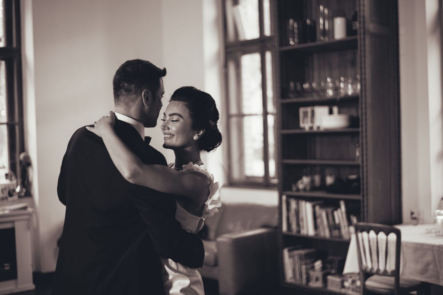 Black and white photo of bride and groom dancing closely in elegant room with bookshelves.