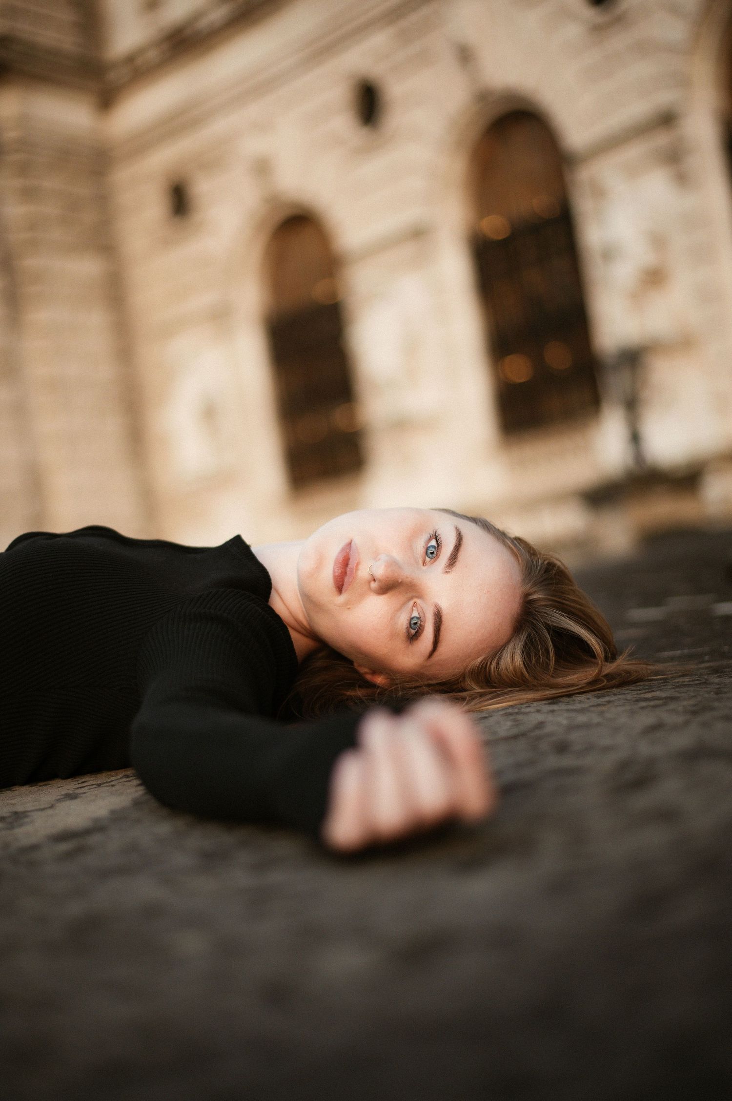 Young woman in black outfit lies on stone surface looking up at camera near historic arched building.