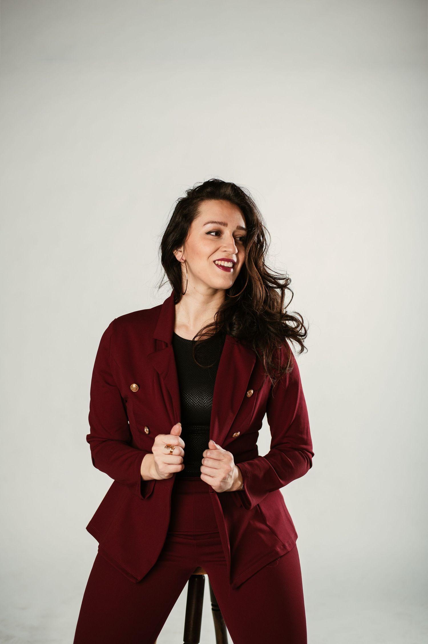 Cheerful brunette woman in a burgundy double-breasted blazer laughs while posing in a white studio.