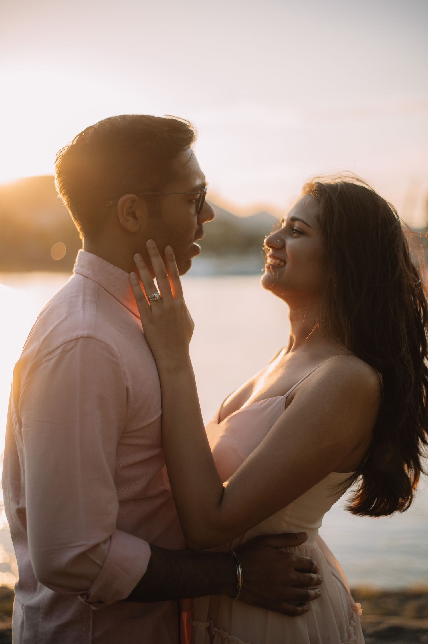 Romantic couple embracing at golden sunset by the water, woman tenderly touching mans face while smiling.