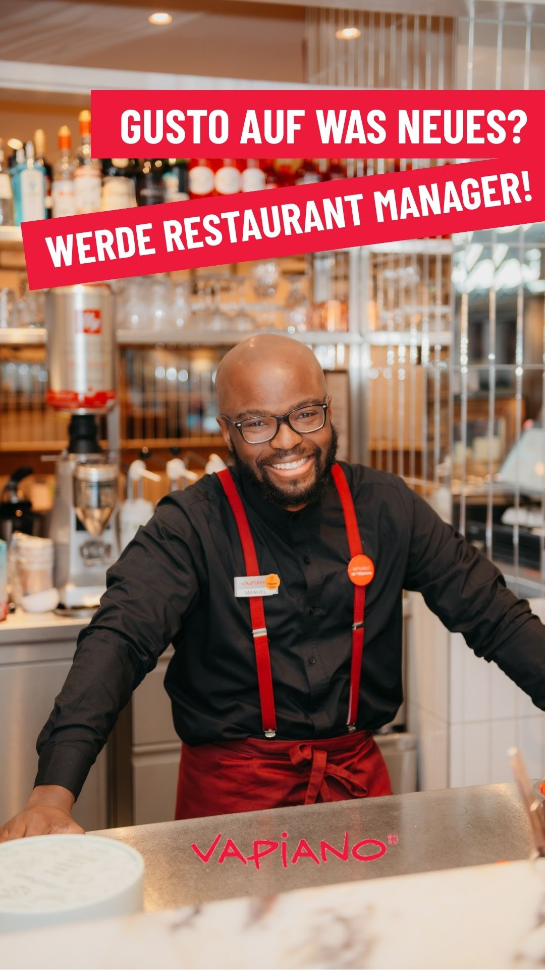 Smiling Vapiano restaurant manager in black shirt and red apron poses at counter with recruitment ad in German.
