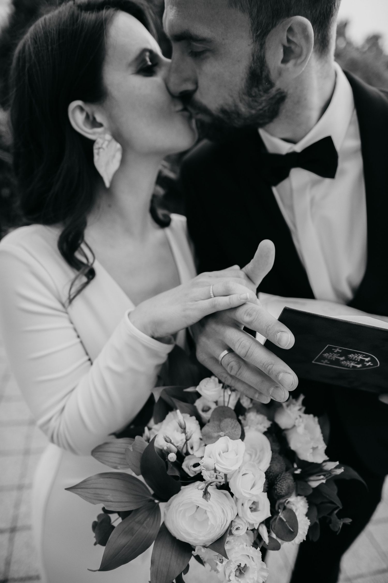 Black and white wedding photo of bride and groom kissing while holding bouquet and bible, showing wedding rings.