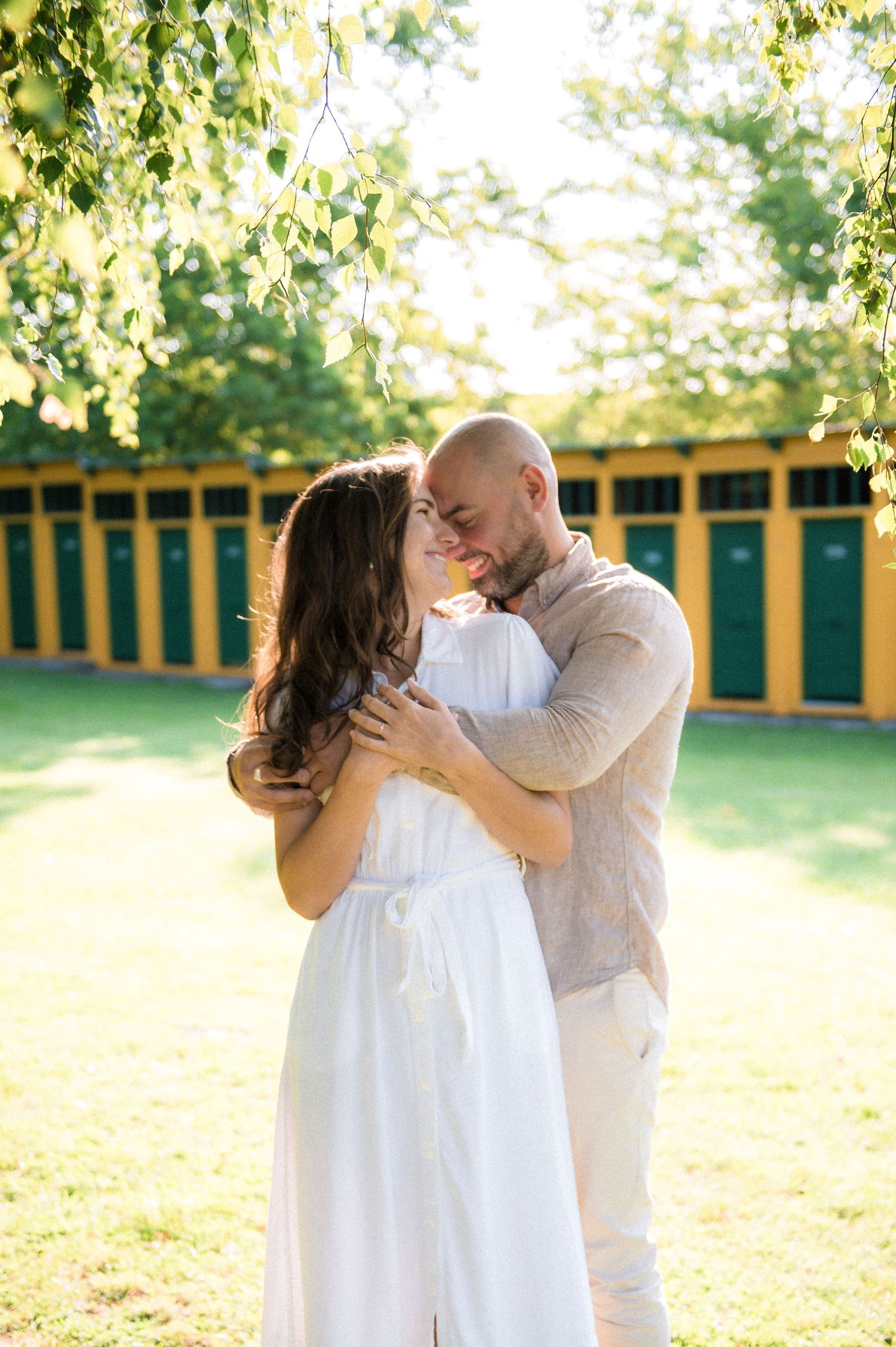 A couple embraces and smiles under birch trees in golden sunlight, woman in white dress, man in linen shirt.
