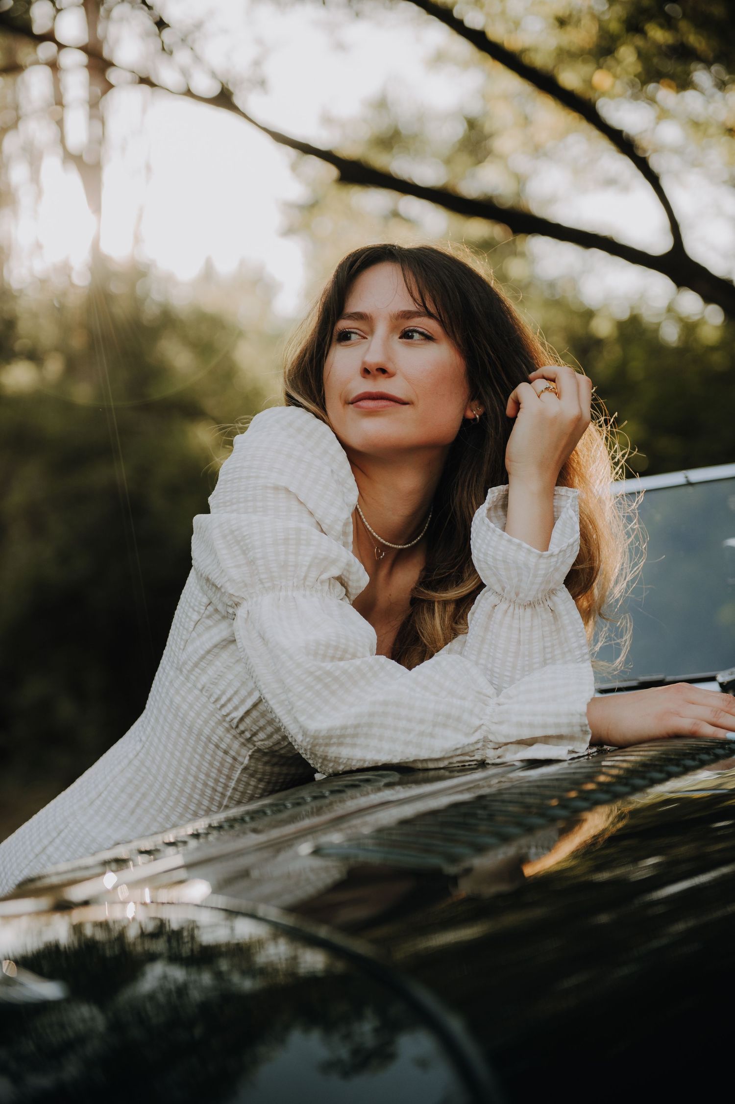 Young woman in white checkered dress leaning on a dark car hood outdoors in golden hour light.