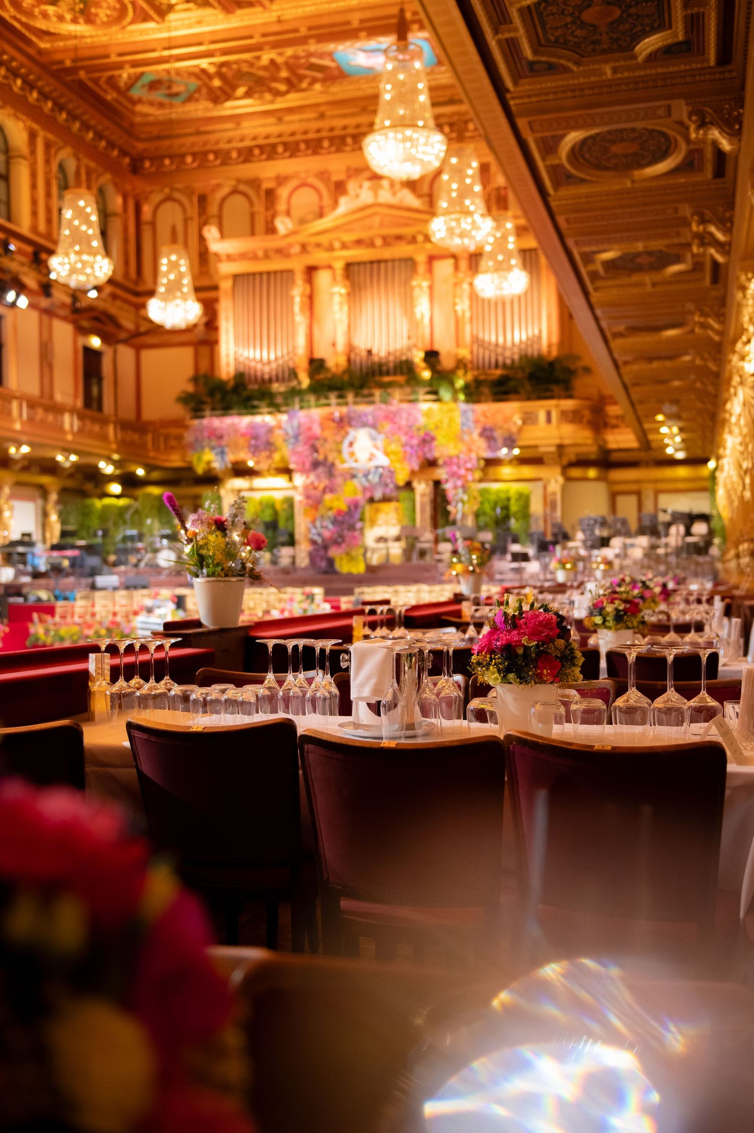 Elegant grand ballroom with ornate gold decor, chandeliers, pipe organ, and tables adorned with colorful floral centerpieces.
