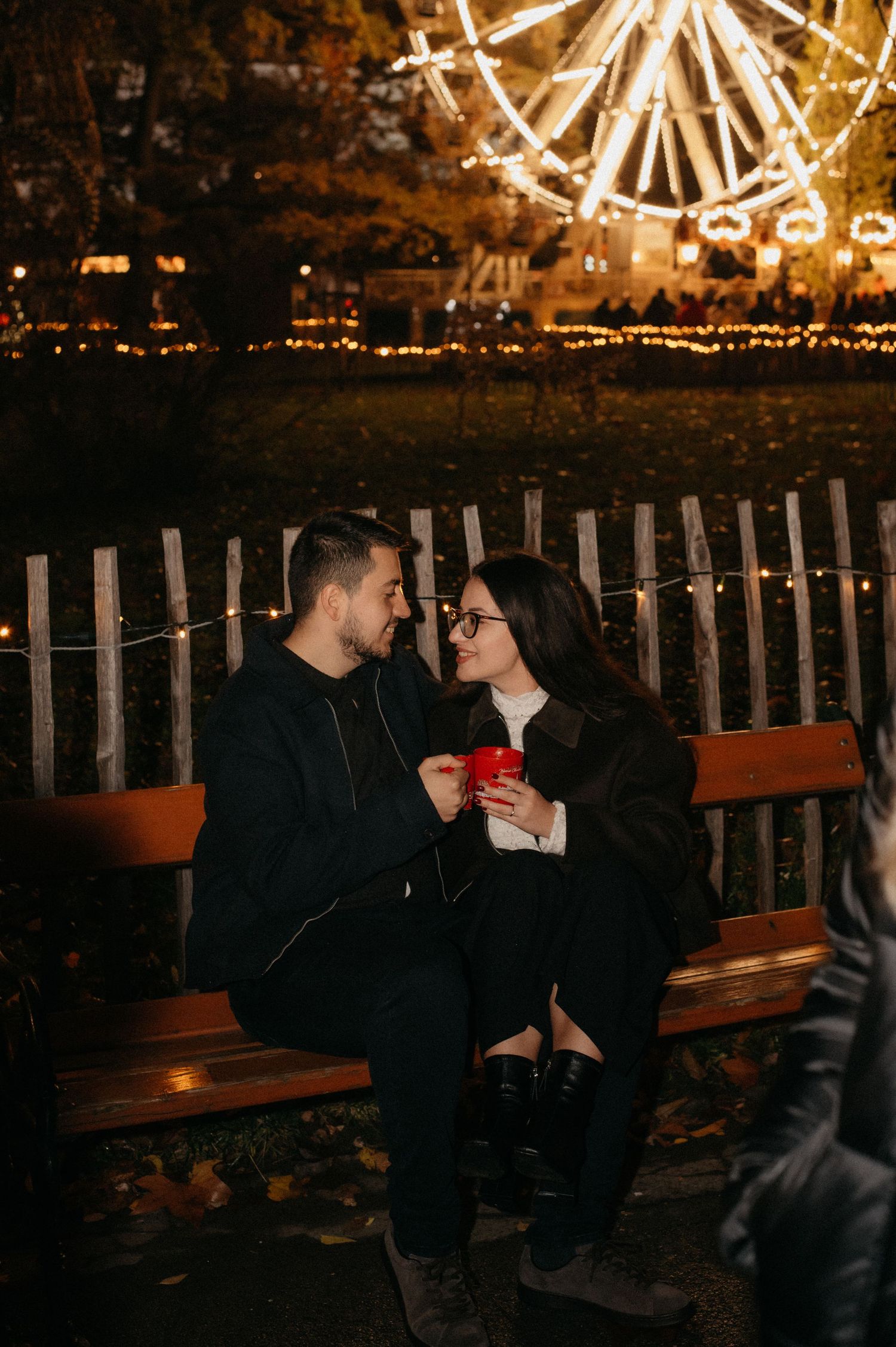 Couple sitting on a bench at night near a lit ferris wheel, sharing a romantic moment with festive lights.