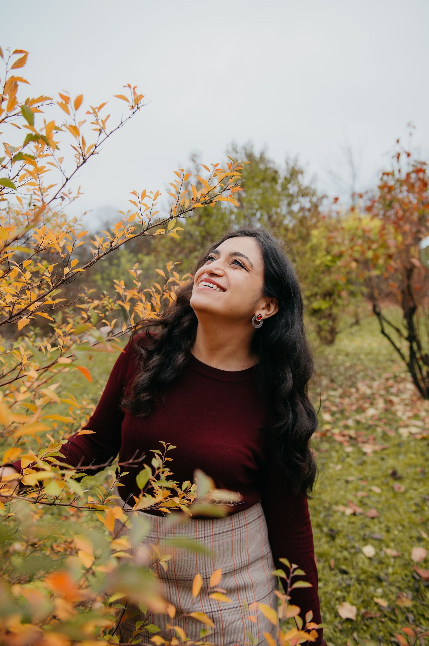 Young woman in burgundy sweater smiling among autumn foliage in an orchard during fall season.